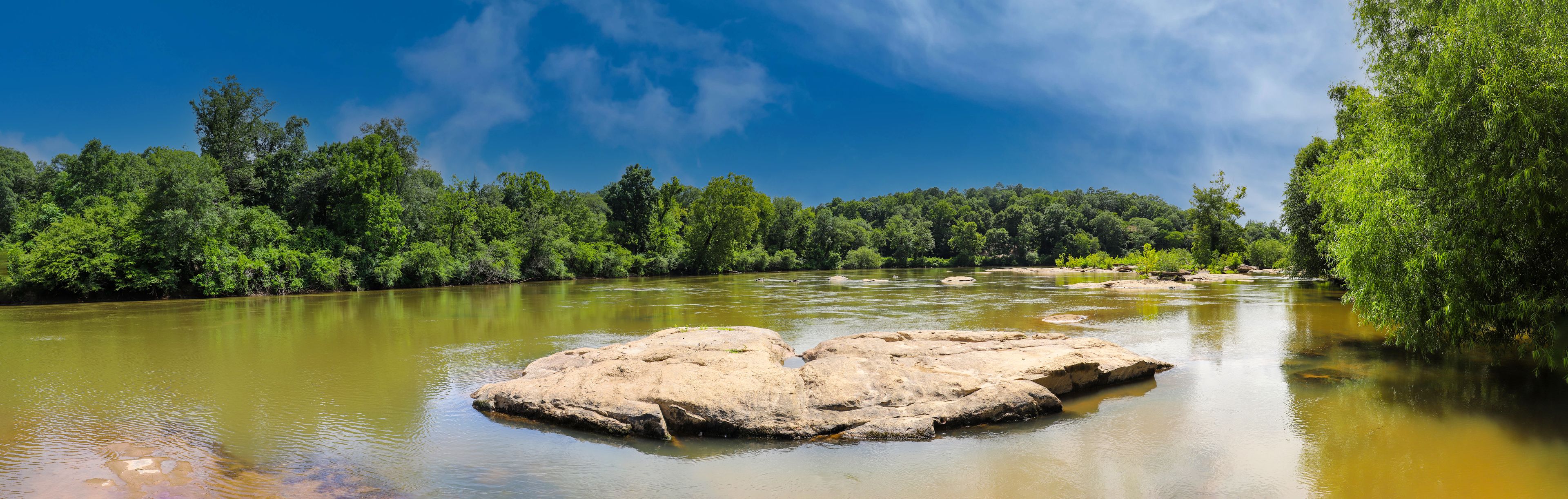 a panoramic shot of the Chattahoochee river with rocks on the middle of the river and green trees reflecting off the water with blue sky and clouds at McIntosh Reserve Park in Whitesburg Georgia