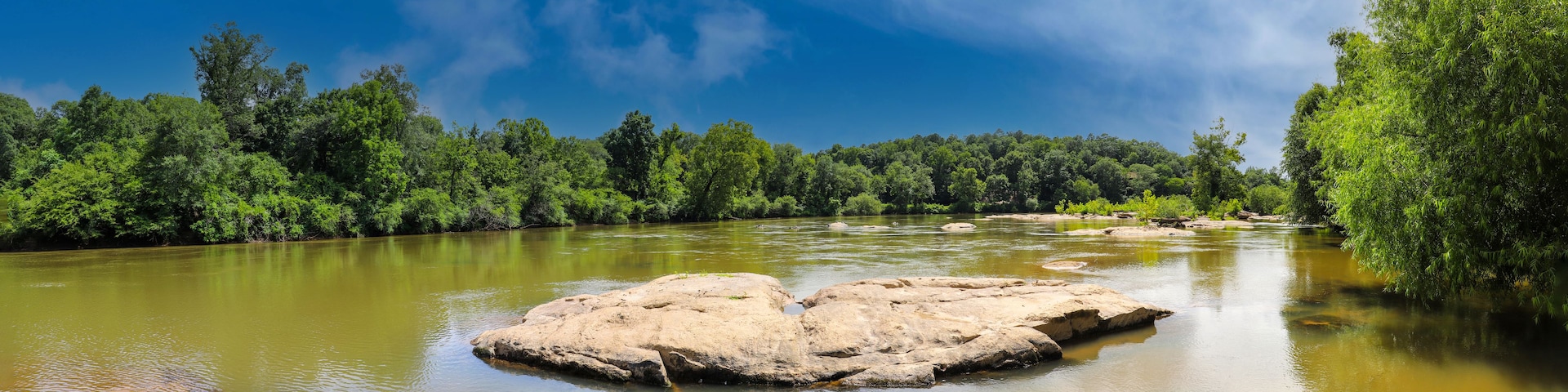 Un río con rocas y árboles verdes frondosos bajo un cielo azul despejado.