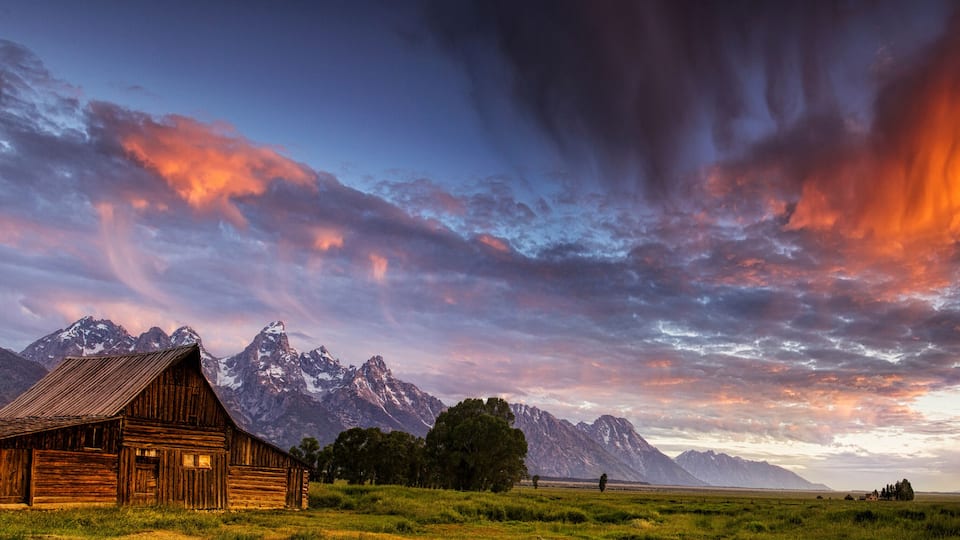 A wooden cabin, green field, and snow-capped mountains under a colorful sky.