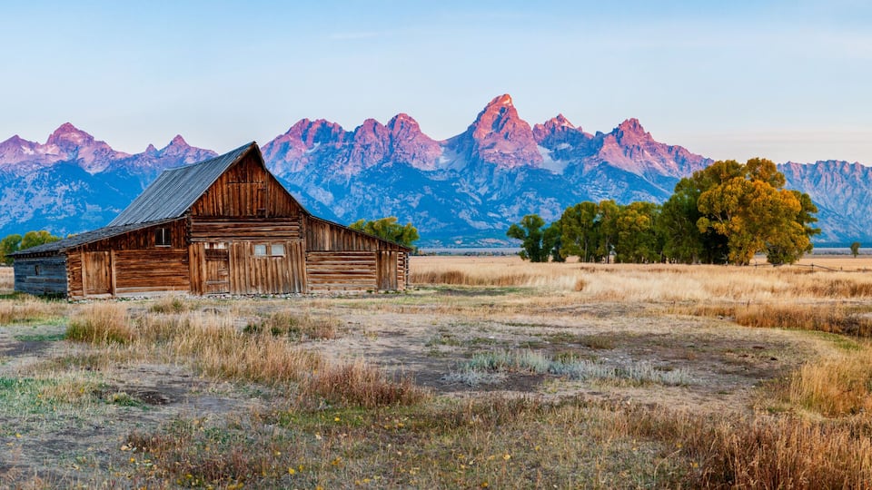 A rustic wooden barn in a field with dry grass, a dirt path, and a mountain range in the background.