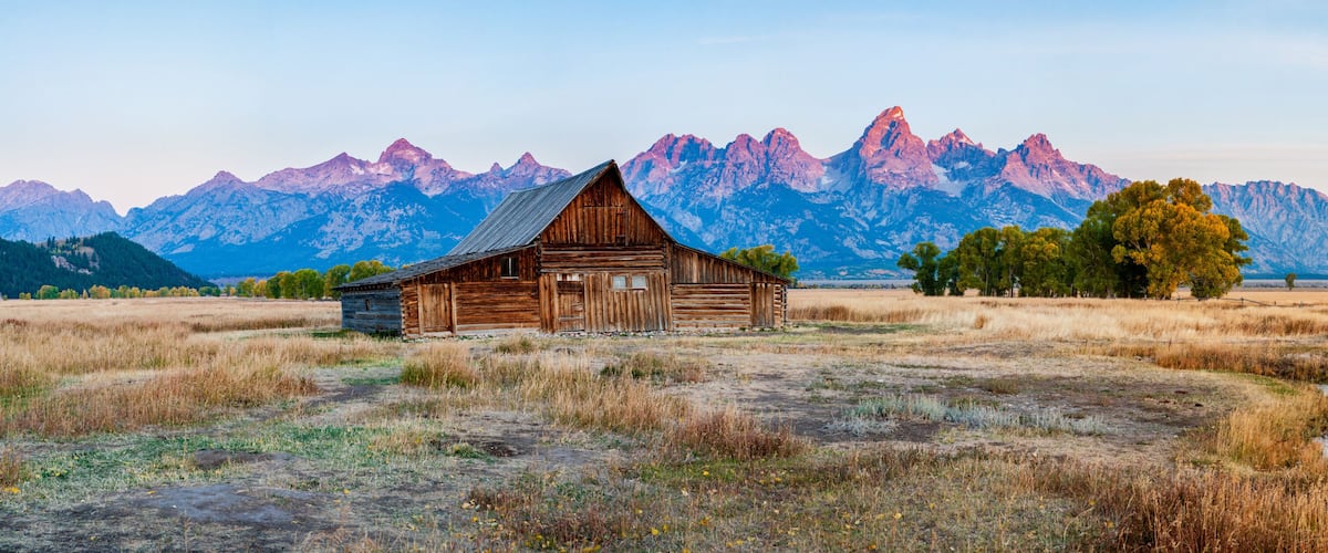 A rustic wooden barn in a field with dry grass, a dirt path, and a mountain range in the background.