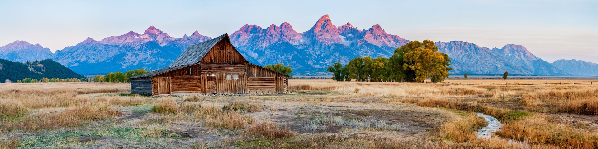 A rustic wooden barn in a field with dry grass, a dirt path, and a mountain range in the background.