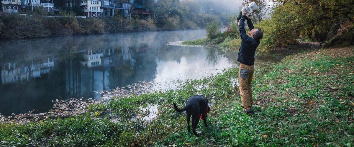 A man is holding a baby near a river and a dog