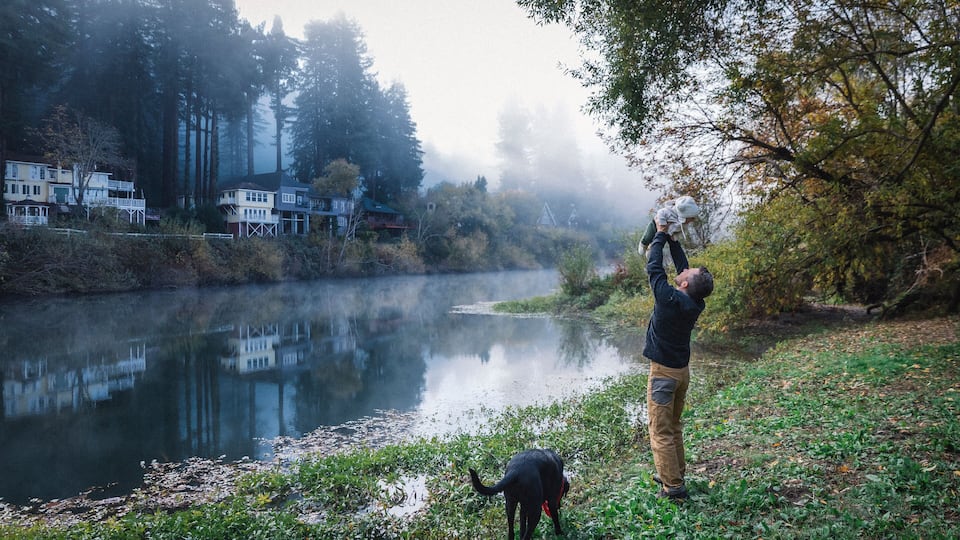 A misty river with houses on the left and a person with a dog on the right.