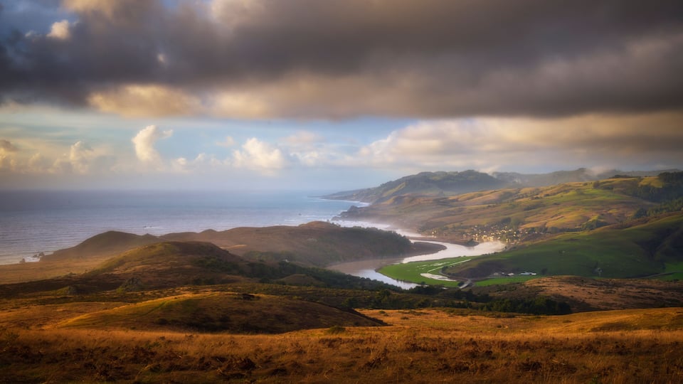 A coastal landscape with rolling hills, a river, and a distant shoreline.