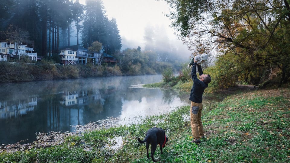 A misty river with houses on the left and a person with a dog on the right.