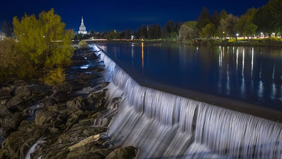 A nighttime waterfall, a calm river, and a distant illuminated tower.