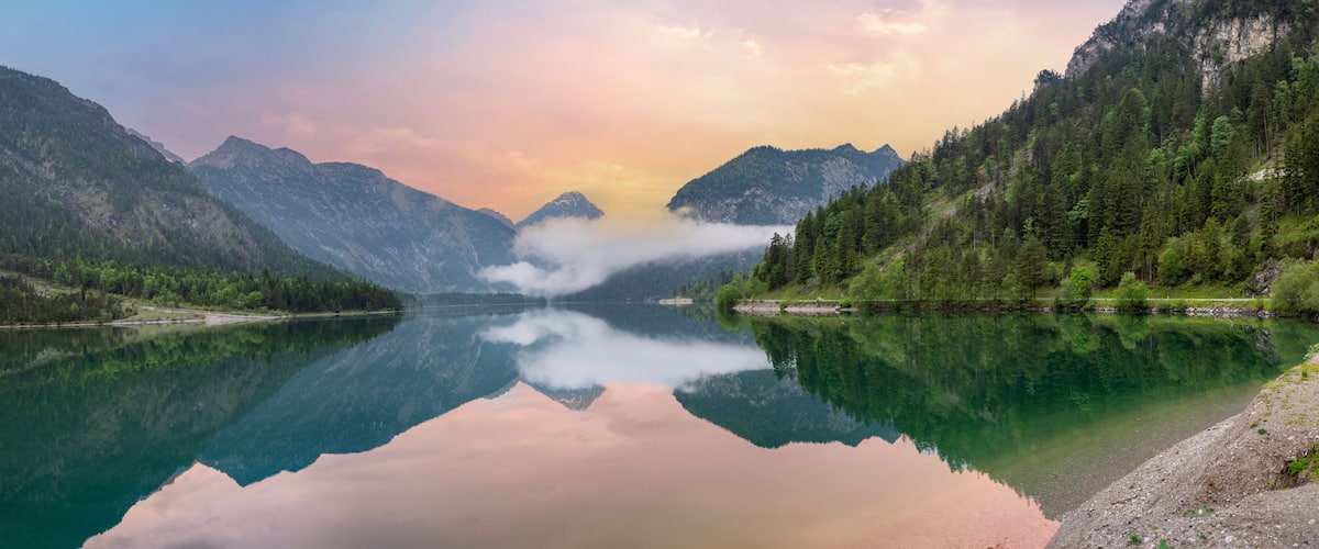 A mountain lake with clear reflections, surrounded by forested slopes and a misty atmosphere.