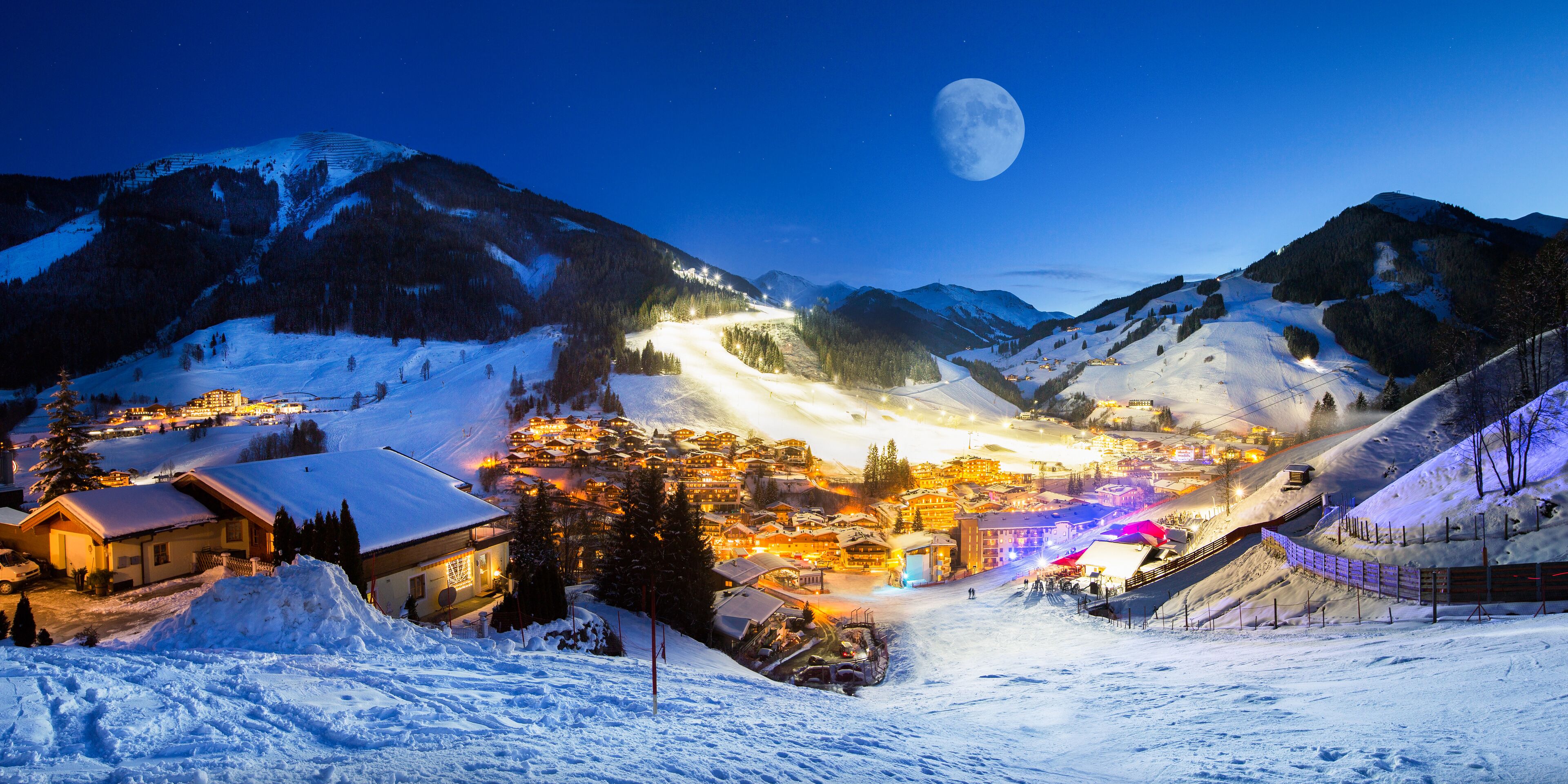 Un pueblo nevado en la montaña, de noche, con una luna llena en el cielo y edificios iluminados.