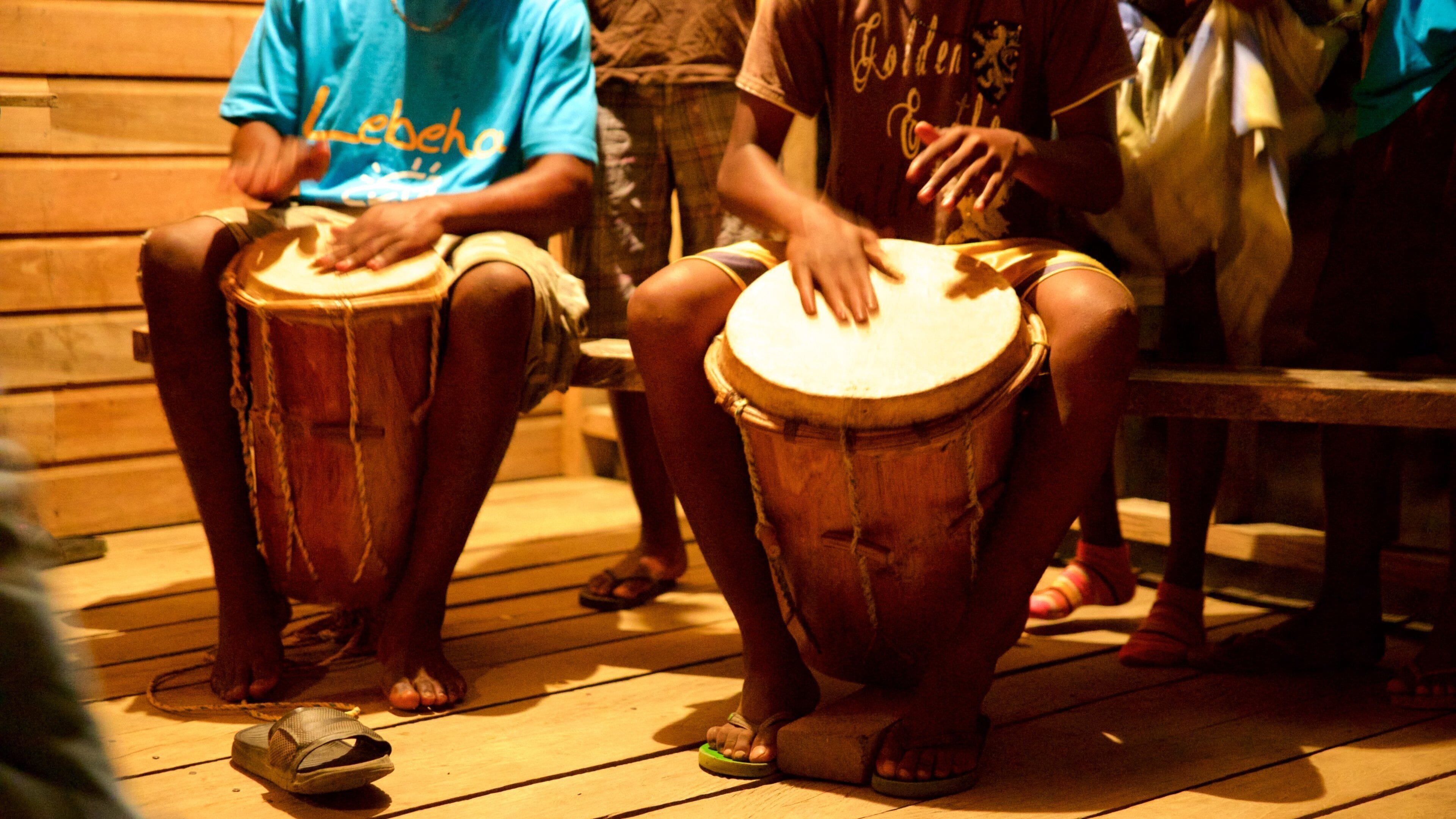 People playing drums in a wooden-floored room.