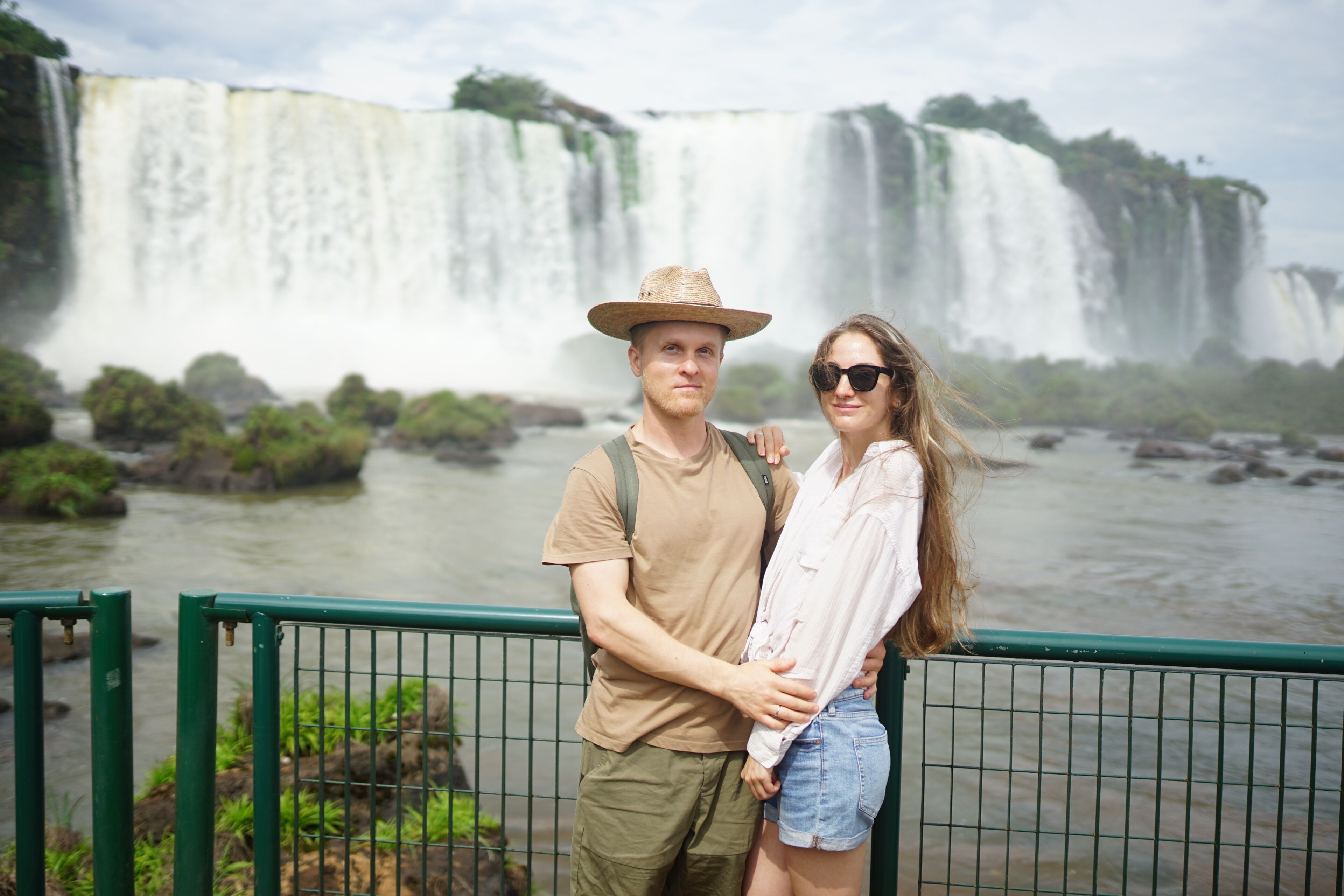 Um casal em frente a uma grande cachoeira, com um rio ao primeiro plano.