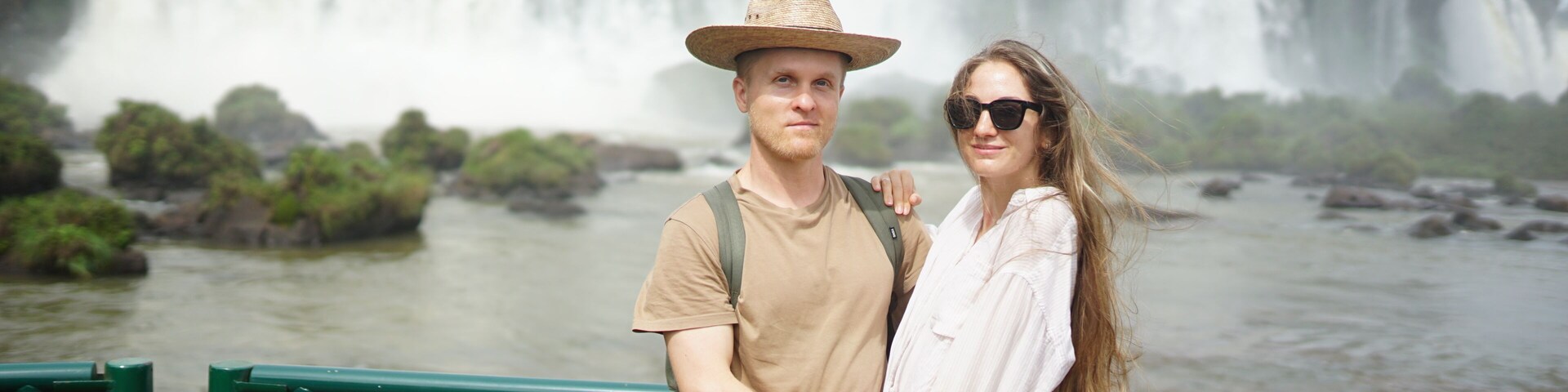 Um casal em frente a uma grande cachoeira, com um rio ao primeiro plano.