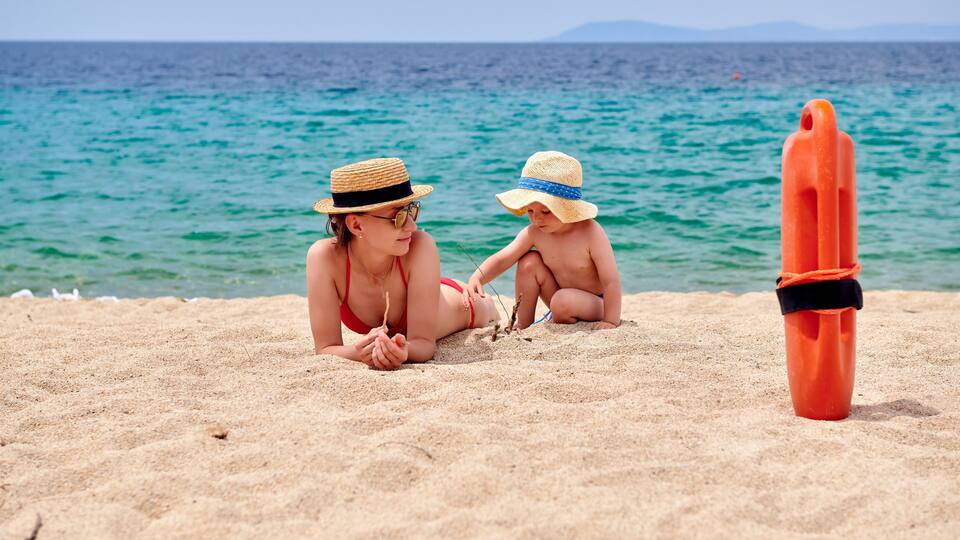 Two people on a sandy beach with a clear blue sea and mountains in the distance.