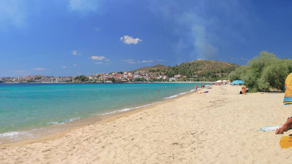 A beach with clear turquoise water, a sandy shore, and a distant town on a hill.