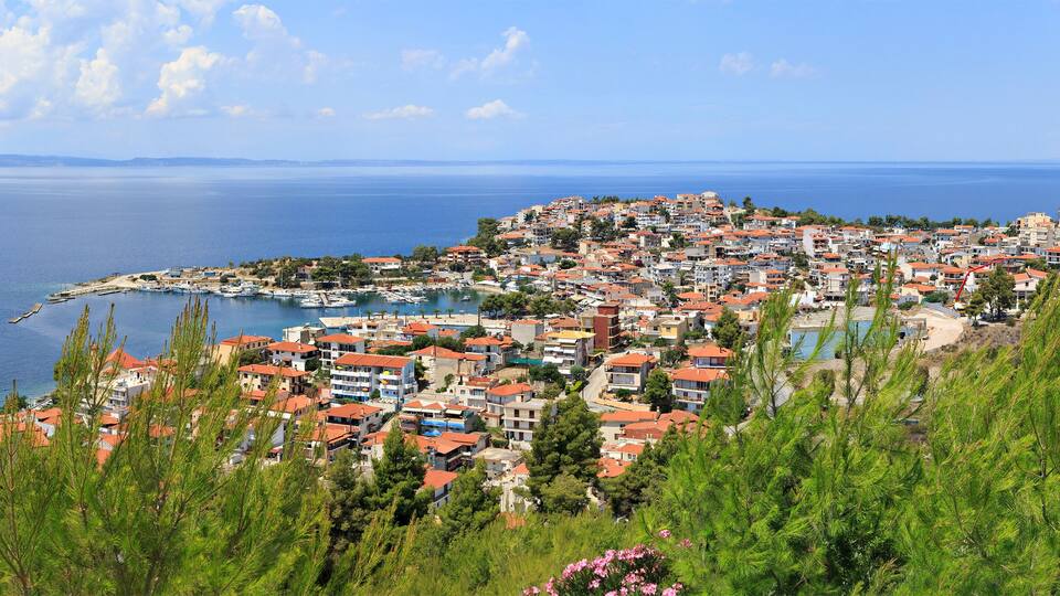 A coastal town with red-roofed buildings, a marina, and a view of the sea and distant islands.