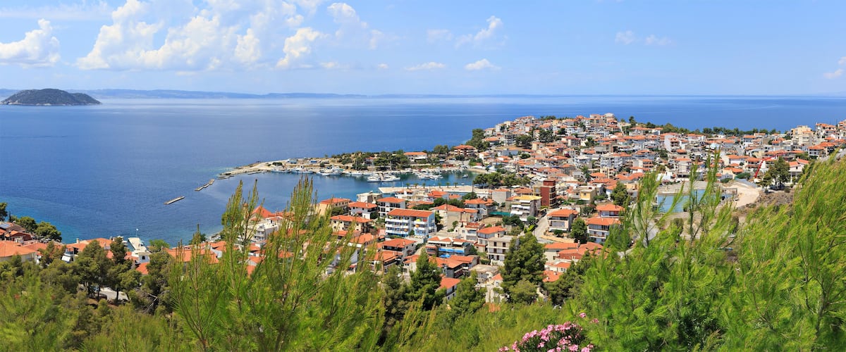 A coastal town with red-roofed buildings, a marina, and a view of the sea and distant islands.