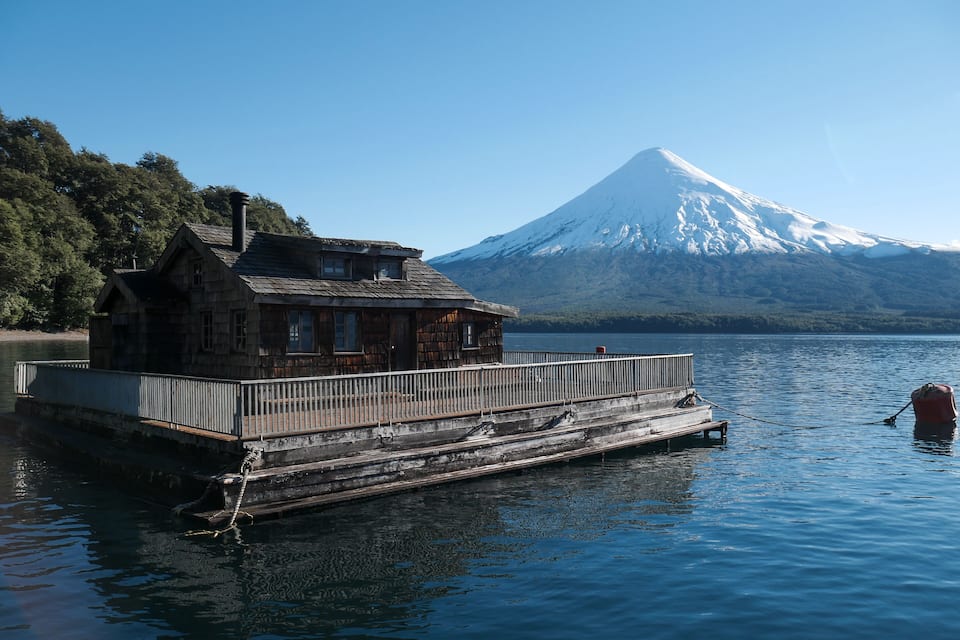 A wooden house by a lake with a snow-capped mountain in the background.