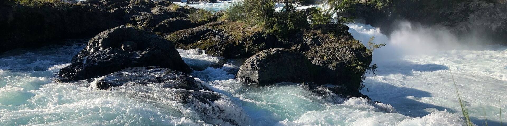 A turbulent river with rocks, surrounded by lush greenery and a mountain in the background.