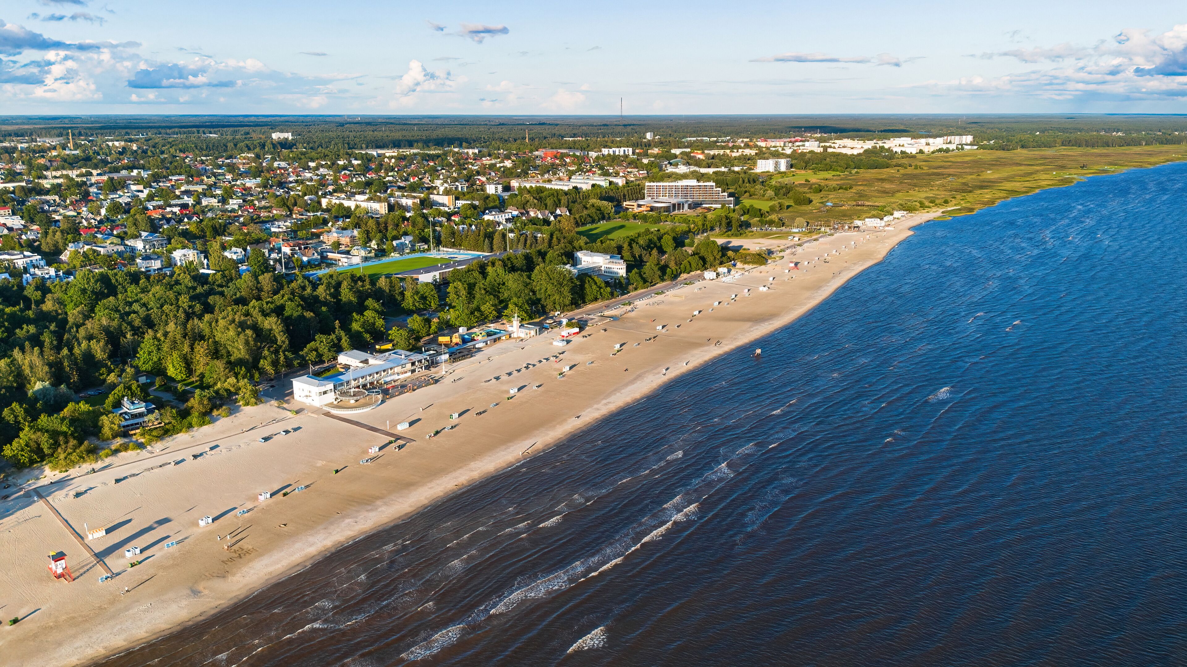An aerial view of a coastal town with a beach, dense forest, and a mix of residential and commercial buildings.