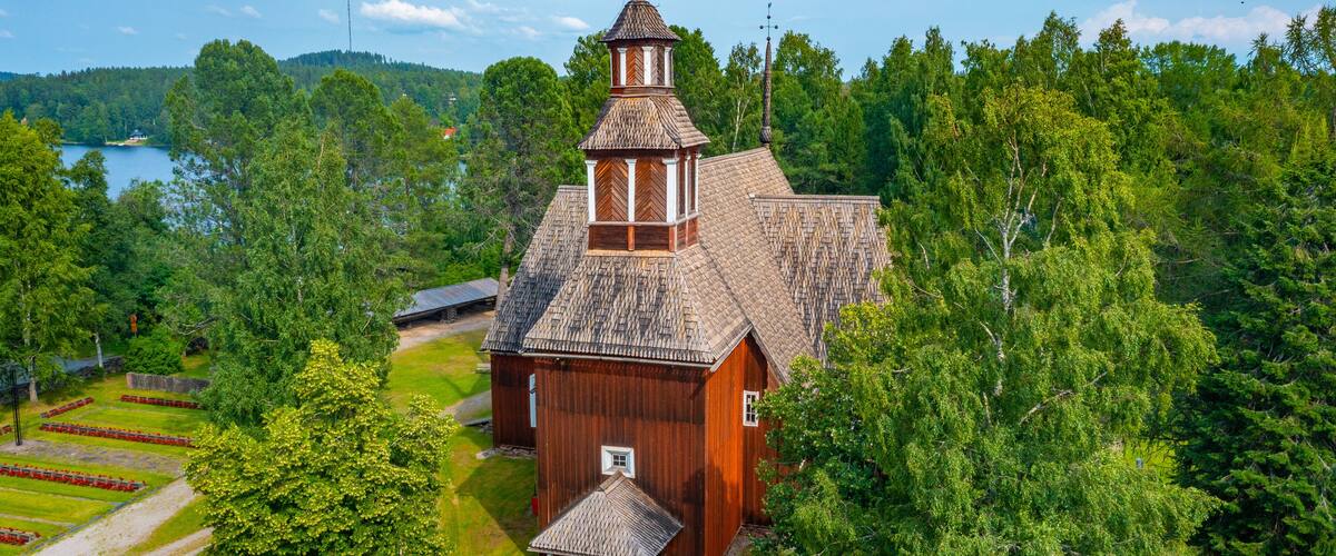 A wooden church with a steeple surrounded by lush greenery and a lake in the background.