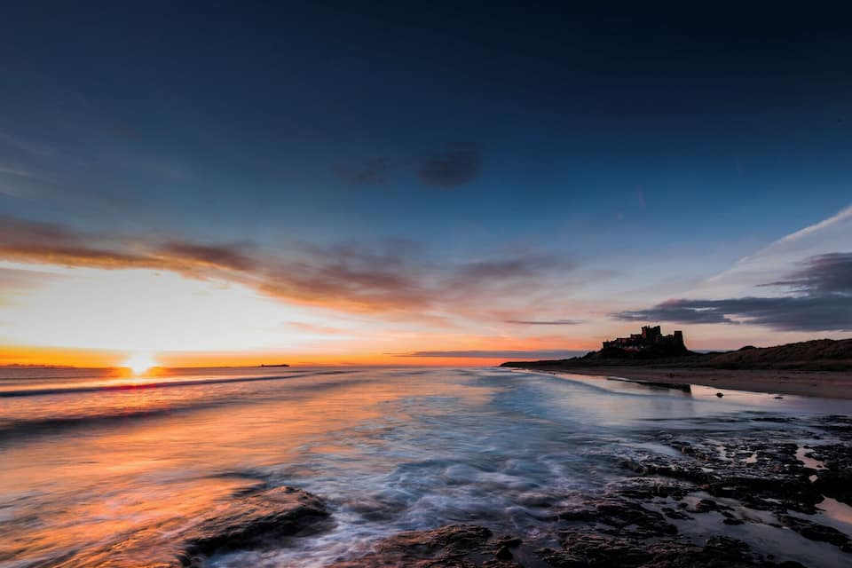 Tramonto su una spiaggia con le onde, un castello in lontananza e cielo sereno.