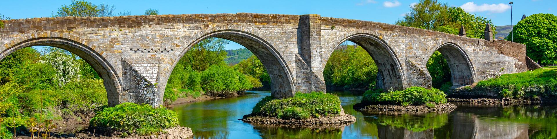 A stone bridge with multiple arches over a calm river, surrounded by lush greenery and trees.