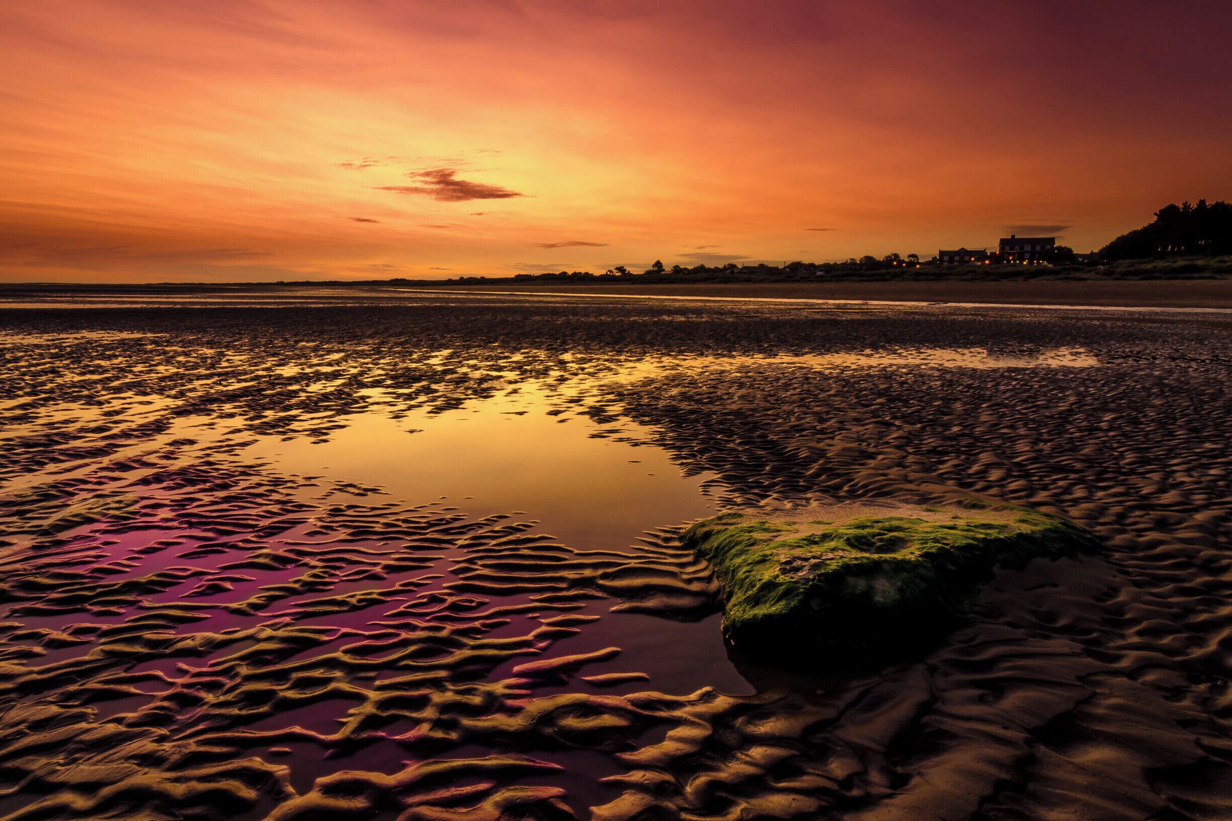 A sunset over a beach with rippled water and a rock covered in moss.