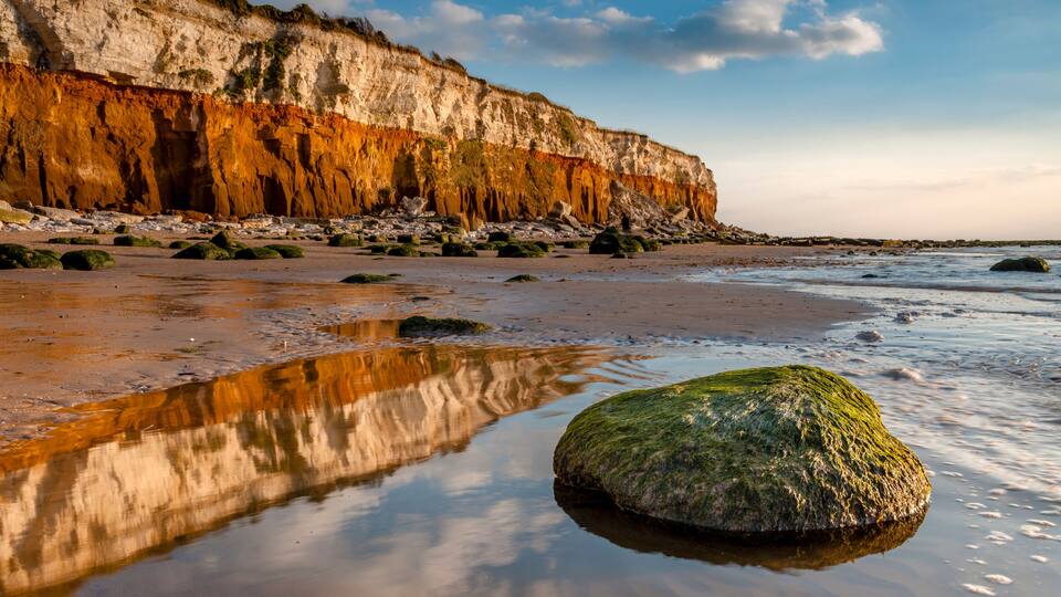 A coastal landscape with a sandy beach, a large rock with green moss, and a cliff with layered rock formations.