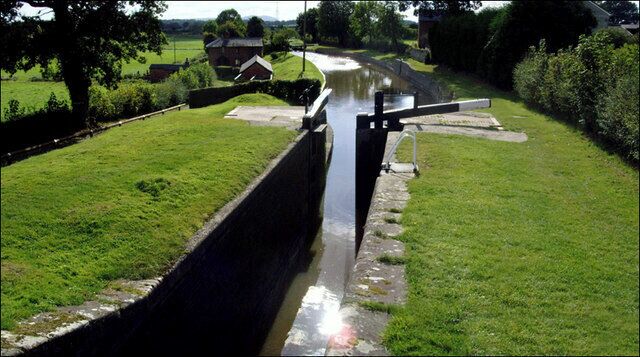 A canal with a lock mechanism, surrounded by grassy banks and a few houses in the background.
