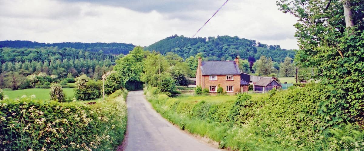 A rural road leading to a house surrounded by lush greenery and hills.