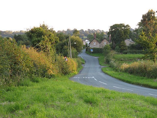 A rural road curving through a green landscape with houses in the distance.