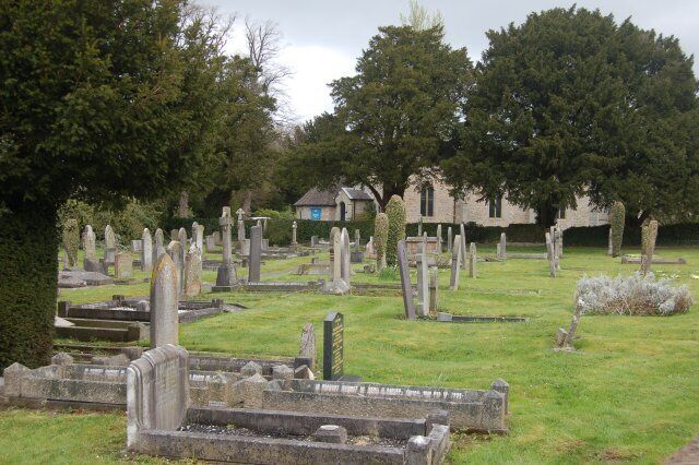 A cemetery with numerous headstones and tombs, surrounded by trees and a building in the background.