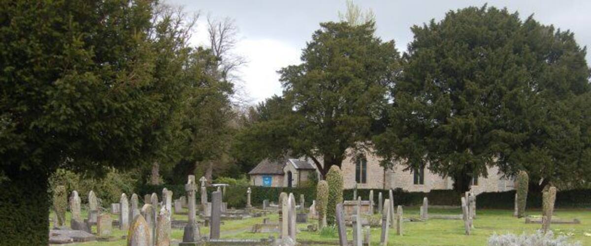 A cemetery with numerous headstones and tombs, surrounded by trees and a building in the background.