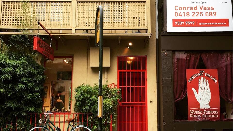 A building with a red door and a balcony. There's a sign for SpaceProperty Estate Agents advertising a 28sqm retail space. A bicycle is parked in front, and there's a sign for 'The Fine Teller' with a hand illustration.