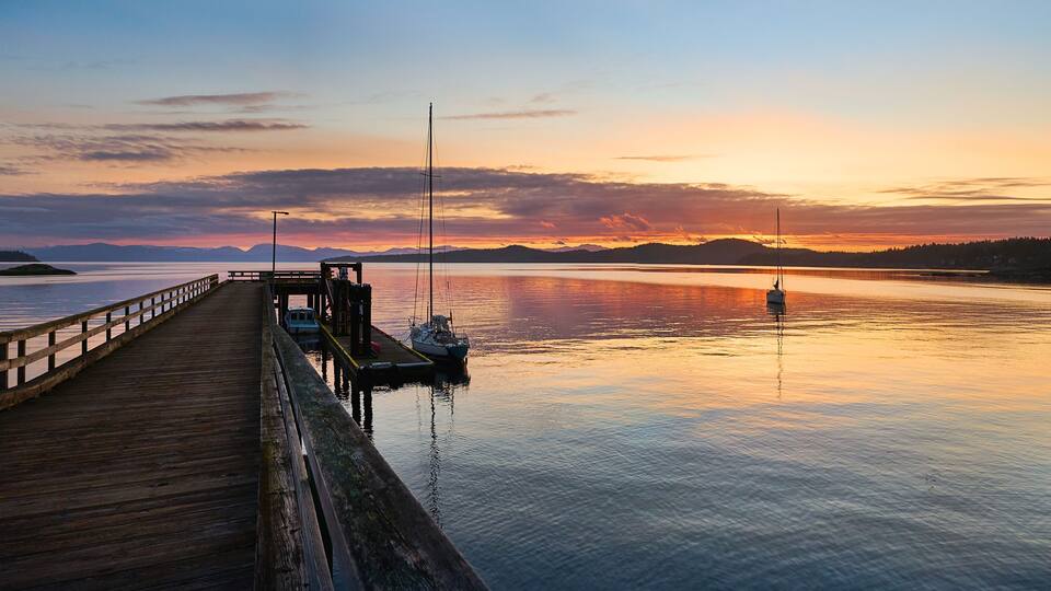 A wooden pier extending into calm waters at sunset with a sailboat docked at the end.