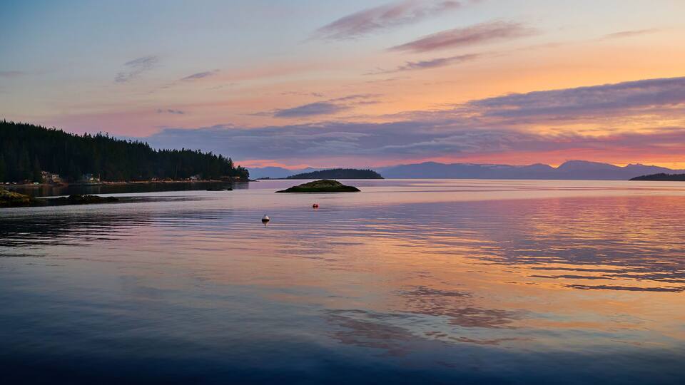 A calm lake at sunset with a forested shoreline and distant mountains.