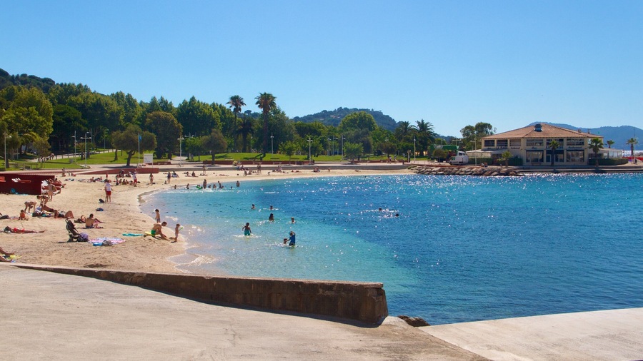 Une plage avec une eau d’un bleu limpide, des gens qui se baignent et qui prennent des bains de soleil, et un bâtiment en arrière-plan.