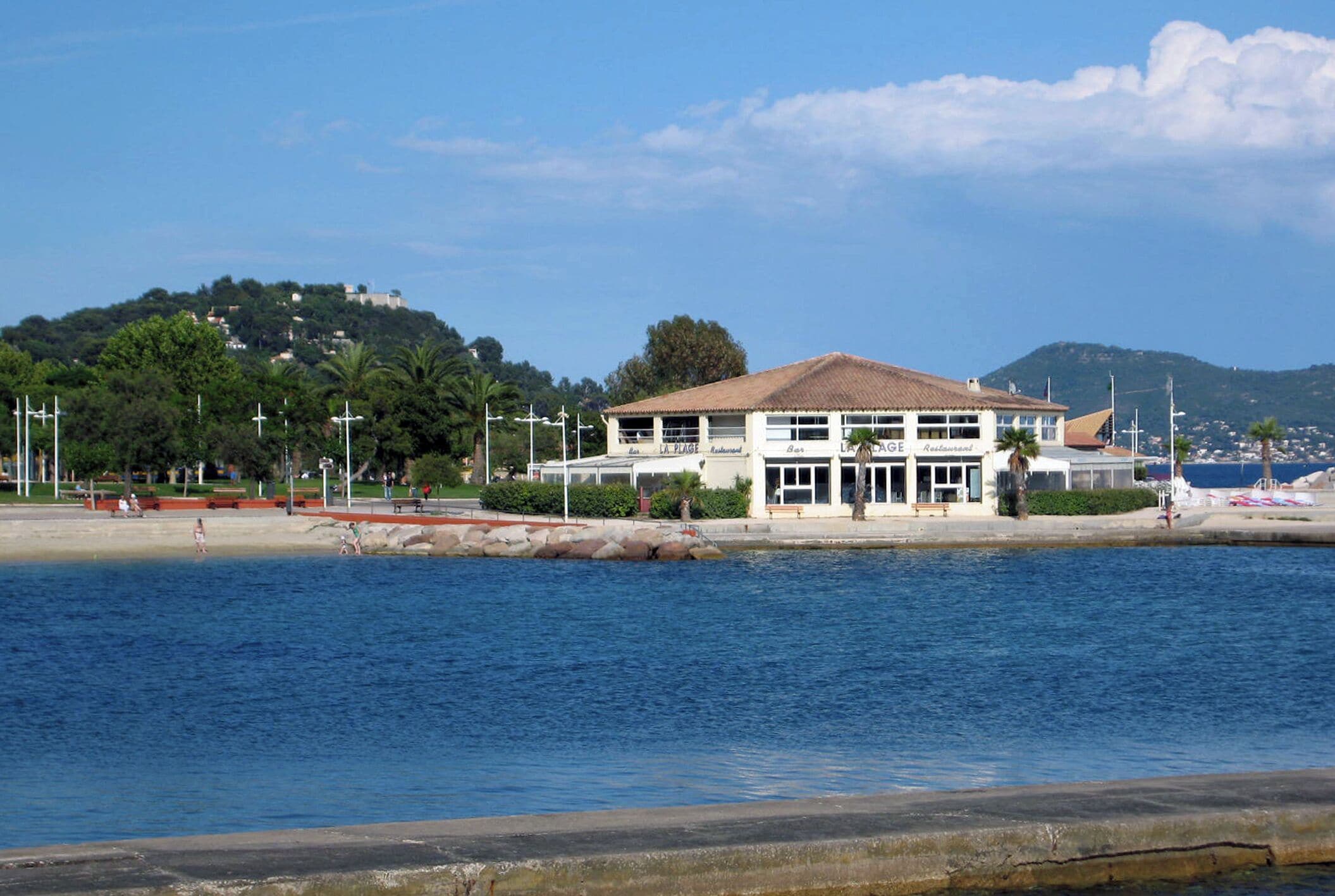Un edificio sul lungomare con tetto in tegole, una spiaggia e una collina sullo sfondo con alberi e edifici.