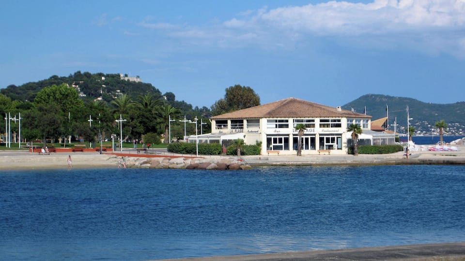 Un edificio sul lungomare con tetto in tegole, una spiaggia e una collina sullo sfondo con alberi e edifici.