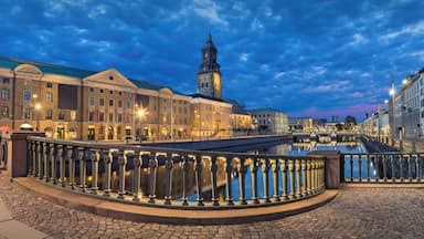 Una plaza histórica en un pueblo europeo, con una imponente torre del reloj, calles empedradas y un puente sobre un canal.