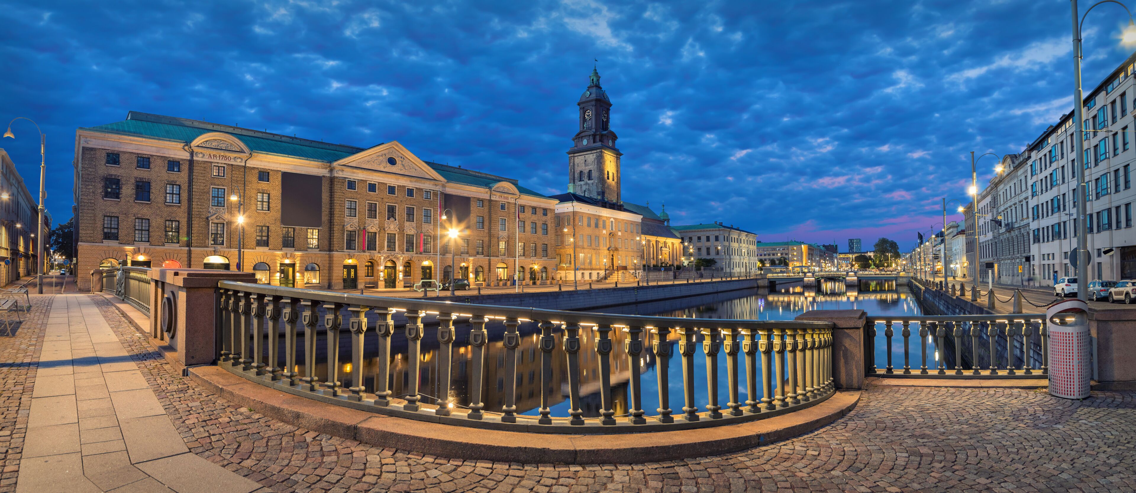A historic European town square with a prominent clock tower, cobblestone streets, and a bridge over a canal.