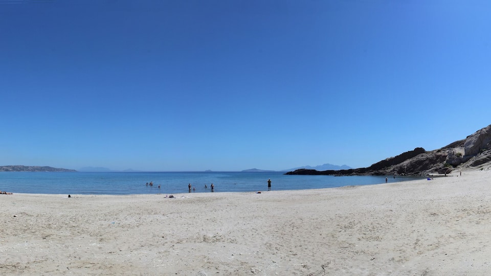 A sandy beach with clear blue water, rocky hills, and distant mountains.