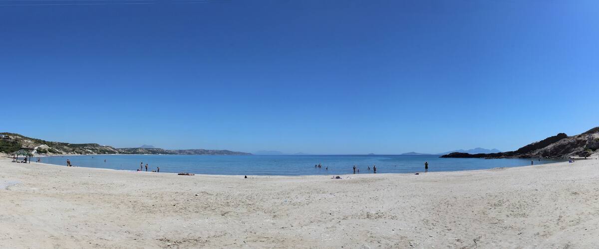 A sandy beach with clear blue water, rocky hills, and distant mountains.