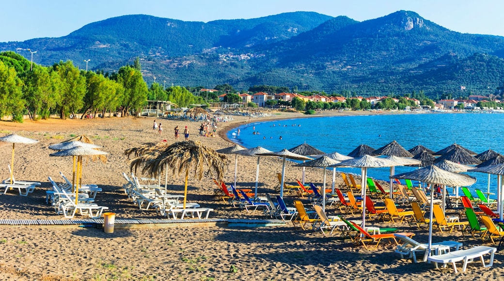 A beach with colorful umbrellas and sun loungers, a calm sea, and mountains in the background.