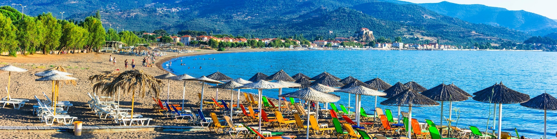 Ein Strand mit bunten Sonnenschirmen und LiegestĂŒhlen, ruhigem Meer und Bergen im Hintergrund.
