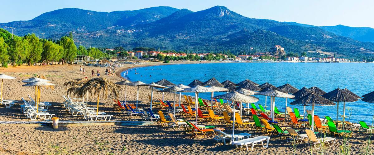 A beach with colorful umbrellas and sun loungers, a calm sea, and mountains in the background.