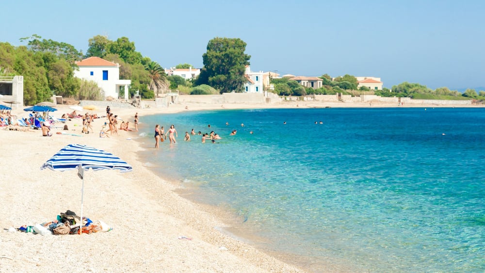 A beach with clear blue water, people sunbathing, and buildings in the background.