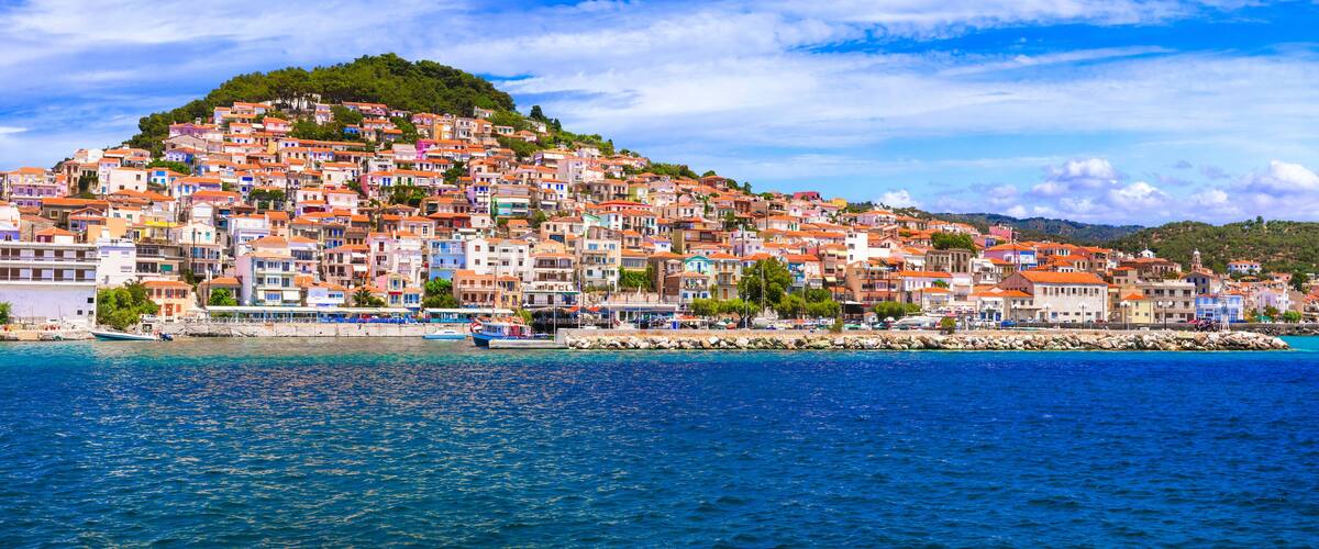 A coastal town with colorful buildings on a hillside, a clear blue sea, and a rocky breakwater.