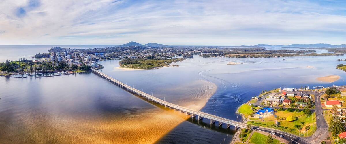 A coastal city with a bridge connecting two landmasses, a large body of water, and distant mountains.