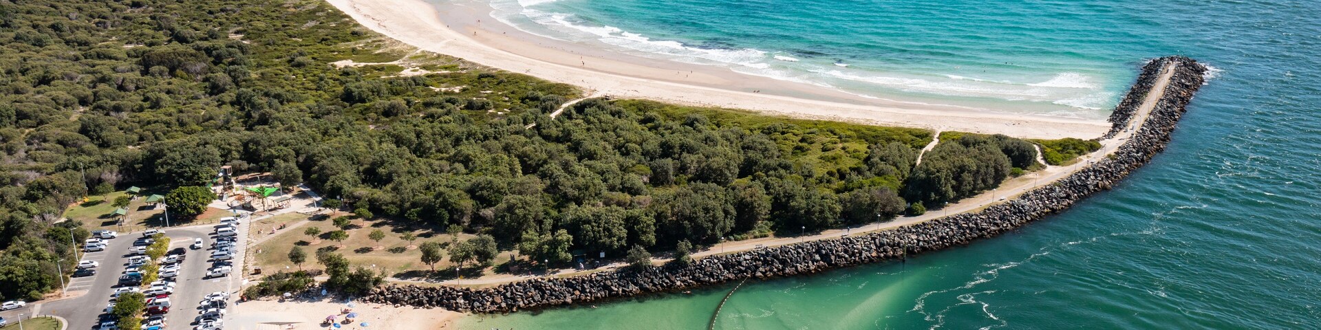 A coastal beach with a curved shoreline, a rocky breakwater, and a parking area.